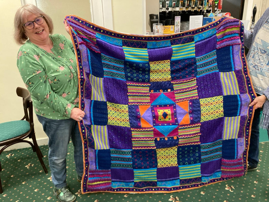 Woman holding up a hand knitted blanket in blues and purple