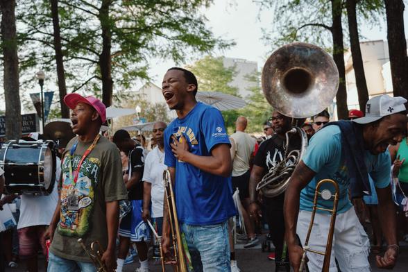 Two men sing with conviction, one holding a brass instrument in one hand with the other hand placed on his chest, a tuba player and crowd in the background