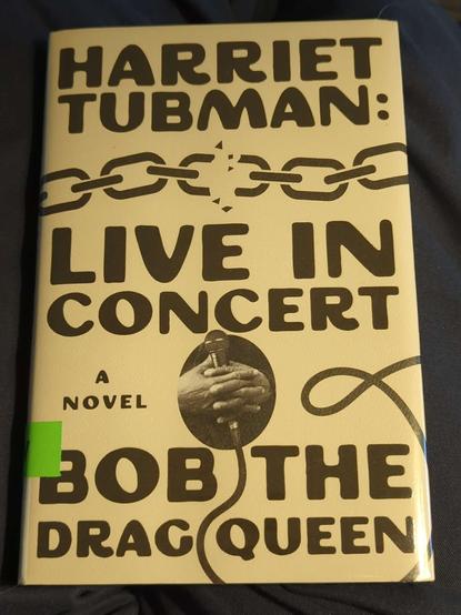 The cover of Harriet Tubman: Live in Concert a novel by Bob the Drag Queen. The background is white, the text a vintage looking black font. At the top is a chain breaking from the center link. In the center between the title and the author name is an oval frame, inside which is a pair of clasped black-skinned hands holding a microphone.