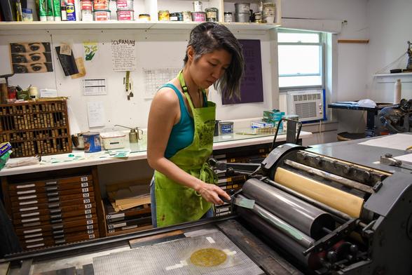 Artist Jenie Gao printing a photopolymer plate on the Vandercook letterpress at Women’s Studio Workshop.
Jenie has short black hair with a side shave and is wearing a teal tank top with a lime green apron.