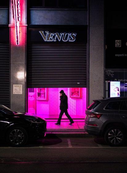 The silhouette of a man walks past the entrance of a night club. Strong magenta light comes from the entrance.