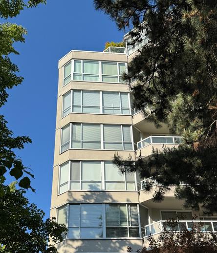 Photo of a residential building showing the top five floors.  The windows are framed in white and all have their white window coverings closed.  To the left and right of the photo are green trees and the sky is clear blue.
