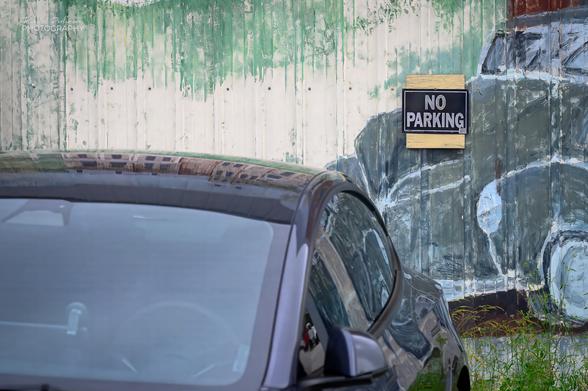 A black car is parked in front of a wall with a mural of a car and a sign that says, "NO PARKING".