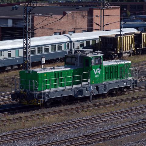 A green VR Group locomotive in a rail yard. Old passenger and coal cars seen on the background.
