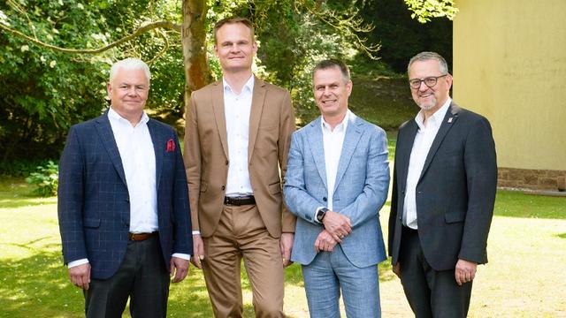 Vier Männer in weißen Hemden und Anzügen sehen in die Kamera. Im Hintergrund ein parkähnliches Gelände und eine Hausmauer.