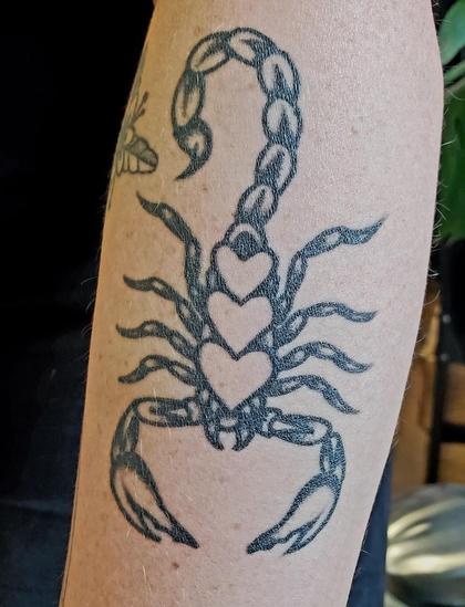 A black ink tattoo of a scorpion is depicted on a fair-skinned arm. The scorpion's body is thick and segmented, leading to a curved stinger. Within the scorpion's main body, there are three small heart shapes. The scorpion has slender legs extended to the sides. Another tattoo of a flower is present.