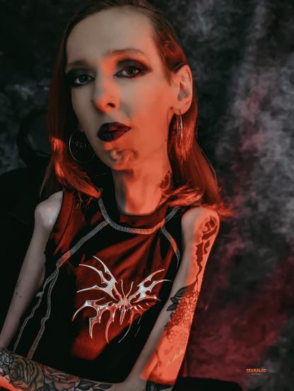 Ein ausdrucksstarkes Porträt von mir. Ich sitze im Rollstuhl, zeige meine auffälligen Tattoos an den Armen und trage ein schwarzes Top mit silbernem Tribal-Motiv. Mein Make-up ist dramatisch: dunkler Lippenstift, intensives Augen-Make-up und betonte Wangenknochen. Rotes Licht fällt über mein Gesicht und meinen Oberkörper und schafft eine düstere, rebellische Atmosphäre. Mein Blick ist ruhig, aber durchdringend – selbstbewusst, trotzig, unangepasst. Im Hintergrund ist ein dunkler, strukturierter Stoffvorhang. Das Bild strahlt Stolz, Stärke und radikale Selbstermächtigung aus.