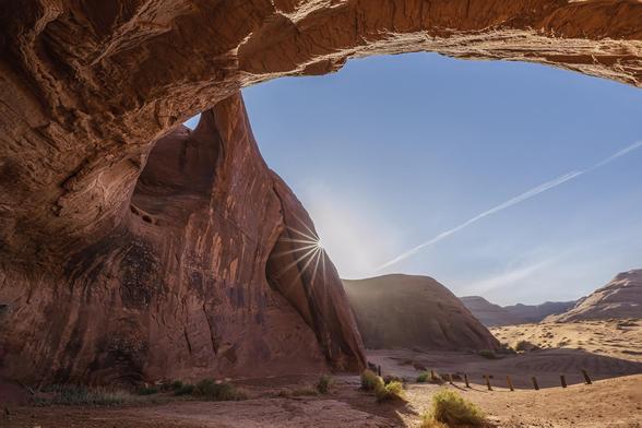 A red sandstone cave on the left of the photo and arching over the top. The early day sun clips the edge of the cave wall creating a sunstar. The Arizona desert is shown in the distance on the right.