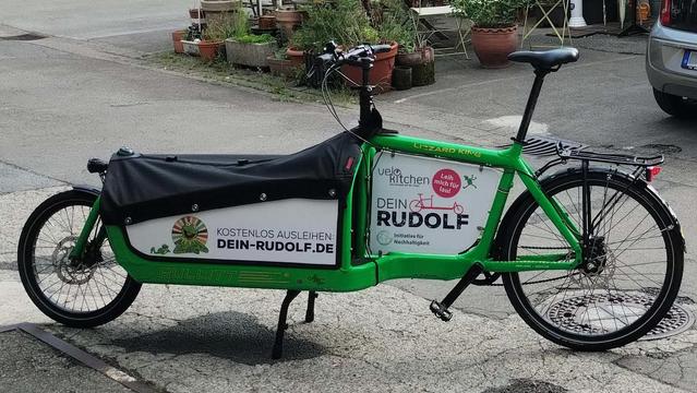 Ein grünes Lastenrad steht auf einer asphaltierten Fläche. Eine weiße Werbetafel im Fahrradrahmen zeigt die Logos von Dein RUDOLF, der Initiative für Nachhaltigkeit und der Velokitchen. Auf der weißen Seitenwand der Ladefläche sind Aufkleber mit dem Frosch Kermit und dem Text "kostenlos ausleihen: dein-rudolf.de"