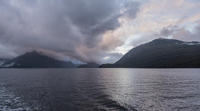 A view from the back of a boat on Doubtful Sound, a fiord in the South West of New Zealand's South Island.  Some of the clouds over the water and hills show a pink tinge, also captured in reflection from the water.  Some bluer sky top right. Some clouds lying over the hills.
