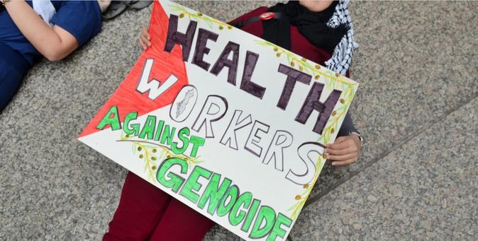 Protestor in a Kefiyeh, laying on the ground, with a sign vaguely in the pattern of the Palestinian flag, that reads: Health Workers Against Genocide.