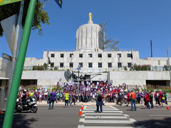 A bunch of union members at a rally outside of the Oregon State Capitol building on a Thursday. The Capitol is a large, brutalist building with a golden statue of a 'Pioneer Man' on the top.