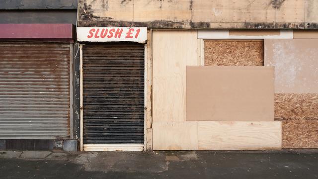 A battered, boarded-up old shopfront. The rusted shutters are down, and the windows covered in a mixture of plywood and OSB. The only hint at the shop's former contents is a sign over the door which reads simply: Slush £1