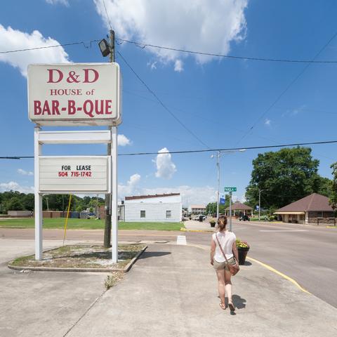 A woman strolls down a fairly soulless and largely empty small-town American street on a hot sunny day. The large sign to her left reads:

D&D House of bar-b-que