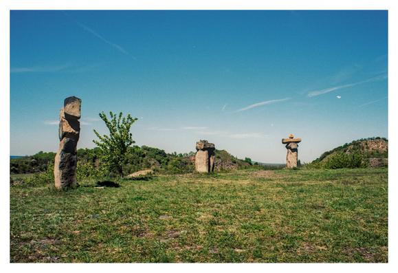 Color photograph of a landscape in Hungary, near Lake Balaton. It shows a meadow-covered plateau on which three stone sculptures stand side by side at a distance, towering into the blue sky.