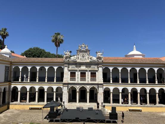 Zu sehen ist die Universität von Evora, Portugal mit Säulen, Bühne im Vordergrund, blauer Himmel und Palmen