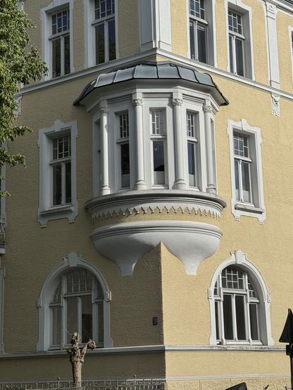Ecke eines mehrgeschossigen Hauses aus der Zeit des Jugendstil, prominent im Vordergrund ist ein fünfteiliges, aufwändig gestaltetes Erkerfenster