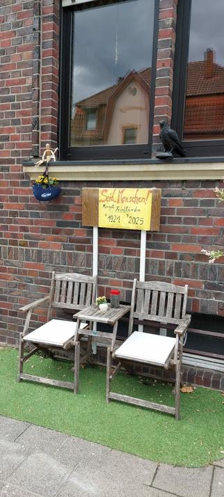 2 Gartenstühle und ein kleiner Tisch aus Holz an einer Hauswand. Darüber ein Schild mit dem berühmten Zitat von Margot Friedländer: Seid Menschen!