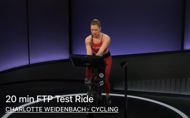 A person is participating in a cycling workout on a stationary bike, focused and engaged. The background features a gradient of purple lighting, and the setup includes a screen displaying the workout title "20 min FTP Test Ride." The individual is wearing a red athletic