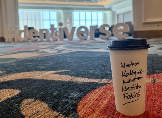 Photo of a coffee cup in front of the Identiverse floor sign, with various names crossed out, ending in “Identity Fabio”.