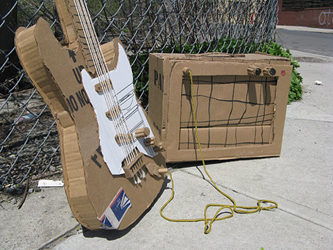Escultura de cartón de una guitarra eléctrica y un monitor en una esquina en la calle