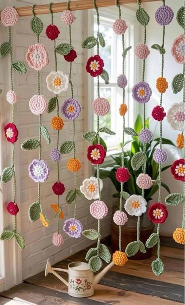 A decorative backdrop featuring colorful crocheted flowers hanging from a wooden rod. The flowers, in various shades of pink, red, purple, and white, are interspersed with green leaves. A watering can sits on the floor beneath the floral arrangement.