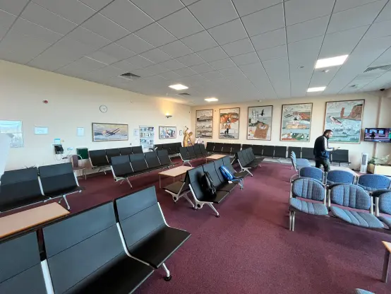 A spacious airport waiting area featuring black and patterned seating, decorative wall art, a clock, and a small television displaying news. One person is standing and checking their phone.
_________

Une salle d'attente spacieuse à l'aéroport avec des sièges noirs et à motifs, de l'art mural décoratif, une horloge et une petite télévision affichant des nouvelles. Une personne est debout et vérifie son téléphone.