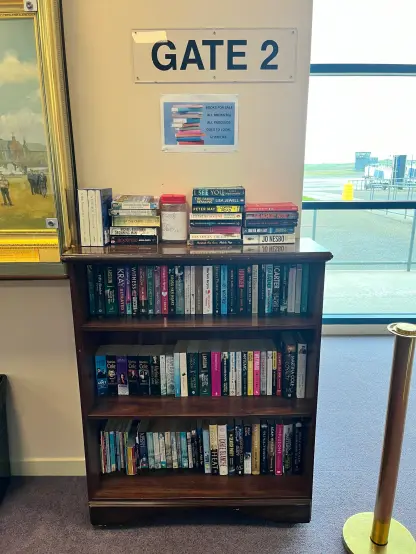 A wooden bookshelf displaying various books for sale, located near an airport gate marked "GATE 2." A sign indicates that all books are priced at 50 pences, with proceeds going to local charities. The background features a window showing an airport view.
___________

Une étagère en bois présentant divers livres à vendre, située près d'une porte d'aéroport marquée "GATE 2". Un panneau indique que tous les livres sont au prix de 50 pences, les recettes allant à des organismes de bienfaisance locaux. L'arrière-plan présente une fenêtre montrant une vue sur l'aéroport.