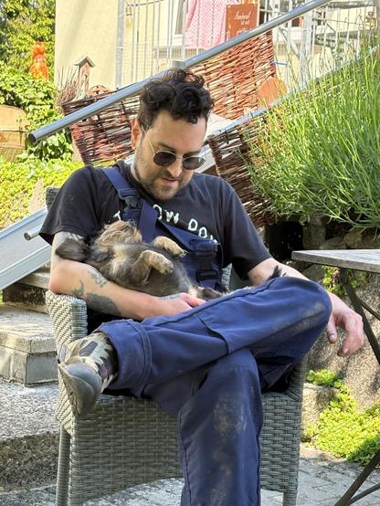 A person sits in a wicker chair outdoors, wearing sunglasses and a black t-shirt while holding a small dog that appears to be relaxed in their lap. The background features greenery and stone steps.