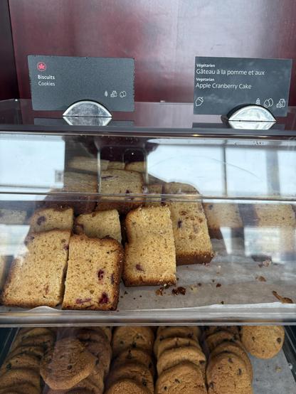 A display with pieces of load cake on the top tier and chocolate chip cookies on the bottom tier. On top, there are two signs in Fetch and English. One says “cookies”, the other one “vegetarian apple cranberry came”
