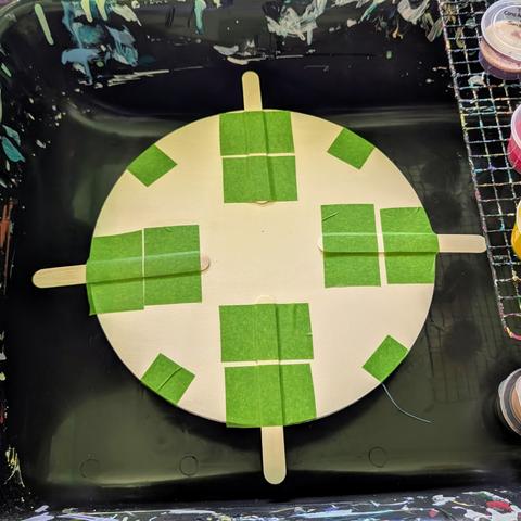 A top view of my paint spinner: there is a circular wooden board with four popsicle sticks taped to it.