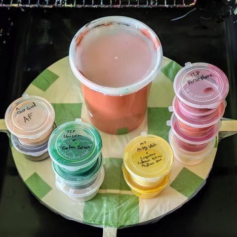 Several containers of paint ready to be used for Shelee bloom and bloom swipe fluid art pours. They're stacked on top of my canvas spinner. The larger container in the back is filled with dark orange house paint, the gold and brown cell activators are on the left, and the other colors are shades of white, green, yellow, and pink.