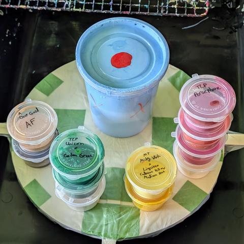 Several containers of paint ready to be used for Shelee bloom and bloom swipe fluid art pours. They're stacked on top of my canvas spinner. The larger container in the back is filled with sky blue house paint, the gold and brown cell activators are on the left, and the other colors are shades of white, green, yellow, and pink.