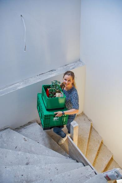 Eine Frau läuft ein Treppenhaus nach oben, trägt Umzugskisten und lächelt dabei in die Kamera.