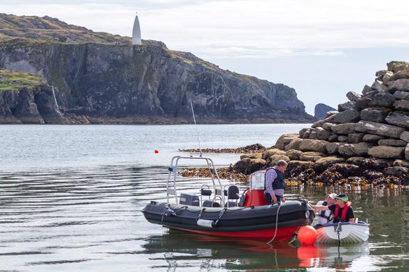 A scenic coastal image taken on Sherkin Island, County Cork, Ireland, showing a small inflatable motorboat and a white dinghy with three people wearing life jackets near a rocky shoreline; a dramatic cliff rises in the background with the distinctive white Baltimore Beacon standing atop it, under a partly cloudy sky.