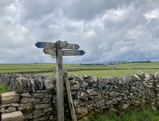 Photo of a six fingered wooden finger post standing next to a dry stone wall. In the background behind the wall are green fields divided up by more drystone walls. In the distance is a farm and behind that the peak of a hill. The sky is full of moody grey clouds.