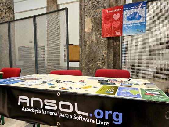 mesa com duas cadeiras e banner a dizer ANSOL - Associação Nacional para o Software Livre e bastantes panfletos e autocolantes sobre software livre