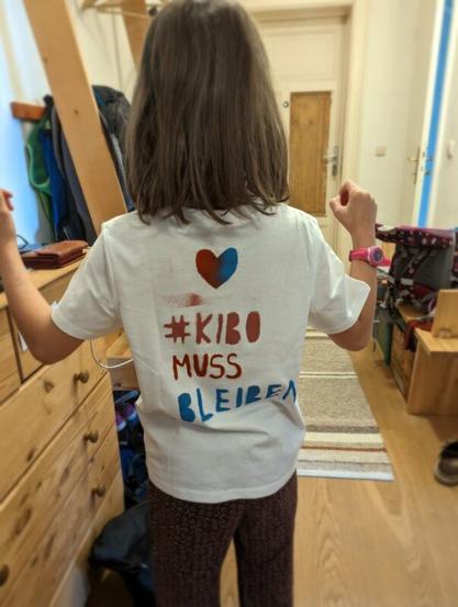 Ein Kind mit einem Demo-T-Shirt von hinten fotografiert. Auf dem Shirt steht "#KIBO MUSS BLEIBEN"