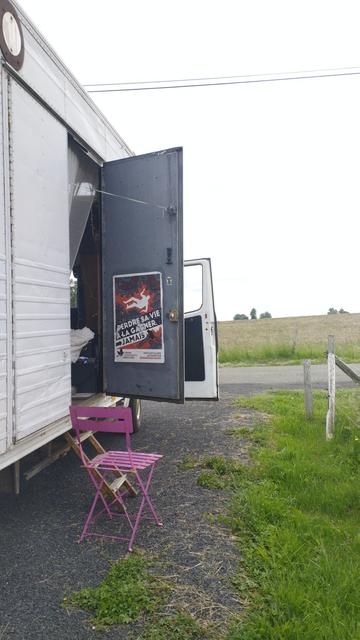 Côté droit du TawaMusi pris de 3/4 arrière, portes de côté ouverte, une chaise rose en métal à côté de la caisse, du gravier au sol, de l'herbe à côté, la route devant une affiche de l'ucl sur la face intérieure de la porte avec le slogan "perdre sa vie à la gagner, jamais"
