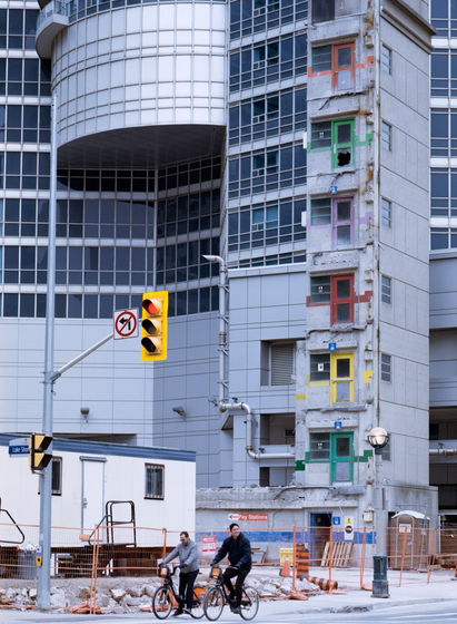 Coloured coded doors mark the floors of the nail-like structure of a parking garage stairwell, left behind as the rest of the garage has been torn down. Cyclists stop and wait at the traffic light below.