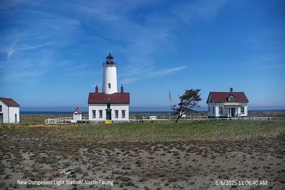 New Dungeness Light Station and Dungeness National Wildlife Refuge today. Located near Sequim, Washington, this image shows several light station structures including the lighthouse and the keeper’s quarters as well as the flag flying between them. The sky today is blue with a hint of clouds. Vegetation and soil of the Dungeness Spit appear in the foreground. The line of deep blue in the background is the Strait of Juan de Fuca.