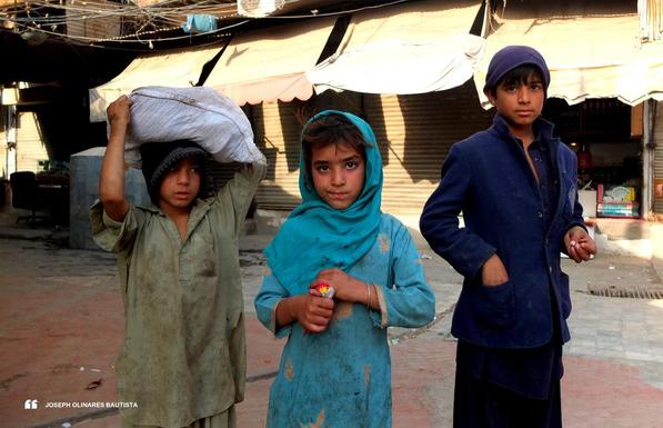 Three children stand side by side on a quiet street in Peshawar, Pakistan, in front of shuttered market stalls. A boy on the left carries a heavy sack on his head, his clothes worn and dusty. In the center, a young girl dressed in a turquoise outfit and headscarf holds a small band of red cloth with gentle determination in her eyes. On the right, an older boy in a deep blue coat and cap stands with confidence, his hand tucked into his pocket. Their expressions are calm, watchful, and quietly strong. Photo taken by Joseph Olinares Bautista in 2013.