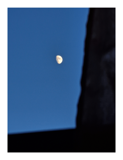 The photo is of a half-moon in a twilight blue sky. A dark, out-of-focus shape, a curtain, is on the right hand side creating a frame. The window sill is on the bottom of the frame and is similarly dark and out of focus. The aim was to create a contrast with the bright moon and clear sky.