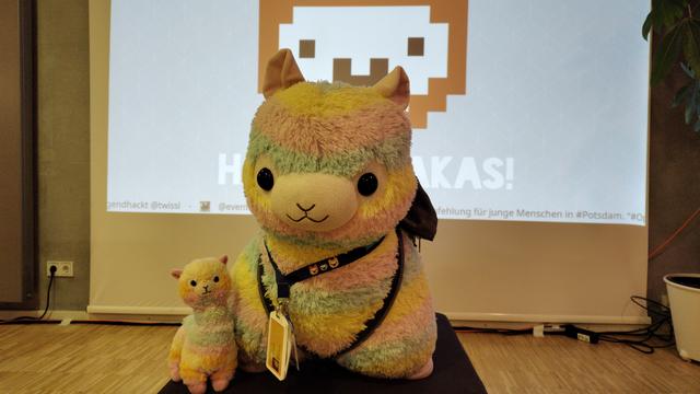 A huge rainbor coloired push alpaka and its smaller sibling sitting in front of a projection screen. Showing a stylized alpaka.