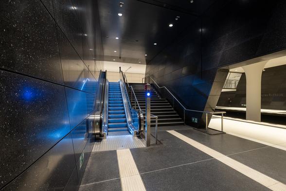 U-Bahnhof Rotes Rathaus in Berlin.
Treppenabstieg zu Bahnsteigebene. Neben der blau beleuchteten Rolltreppe befindet sich eine normal Treppe mit einem Podest.
