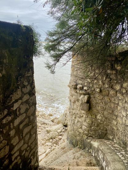 Photo taken from stairs going down to the ocean through a medieval looking lime stone passage. The ocean can be seen between the walls of the passage. Tree branches are decorating the passage to the right.