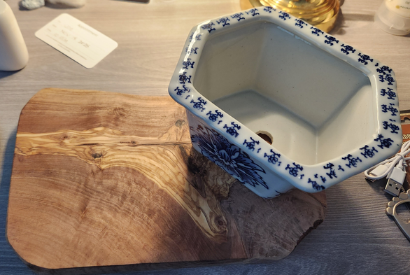 White and blue oblong hexigon pot sitting on a piece of nicely figured olive wood. All sitting on a desk, showing some random things like a business card, base of a brass lamp, and USB cable.