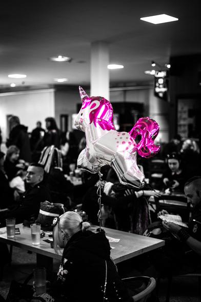 Ein Luftballon-Einhorn mit rosa Mähne und rosa Schwanz schwebt über Tischen, an denen viele Leute sitzen, diese sind schwarz-weiß gehalten.