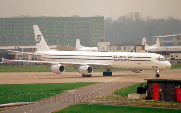 Side view  of a 4 engined jet cargo plane taxiing from left to right.
The plane is mostly white, with a grey belly, and a pair of black stripes running along the body, overlaid with black "Southern Air Transport" text.
The registration "N873SJ" is on the lower rear fuselage, with a stylised, fat, grey letter "S" outlined in black.
Green grass fills most of the foreground, with a grey roadway cutting through, next to a low, black brick building on the right of the frame.
In the background, a collection of white or grey and blue airliners are parked on the right, with a green, metal, box-like structure towering over everything on the left, with trees and grey sky filling the rest of the frame.
