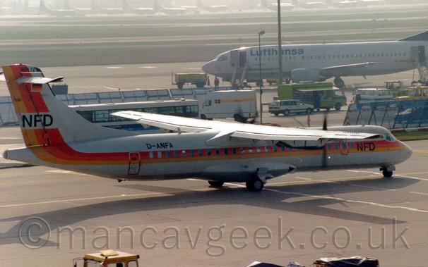 Side view of a high-winged, twin propellor-engined airliner taxiing from left to right.
The plane is mostly white, with a yellow, orange, and red stripe running along the body before sweeping up in to the tail, with black "NFD" letters overlaid under the cockpit, as well as on the tail.
The black registration "D-ANFA"  is on the upper rear fuselage.
Grey concrete apron fills the foreground, with a strong shadow being cast by this plane, suggesting it is sunny.
A collection of airport vehicles are clustered between blue metal fences in the background, with a white, twin engined jet airliner beyond them, with large grassed areas and taxiways beyond that, slowly getting lost in bright haze.